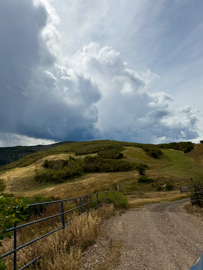 A gravel ranch road beside a metal fence, climbing a green hill under towering storm clouds.
