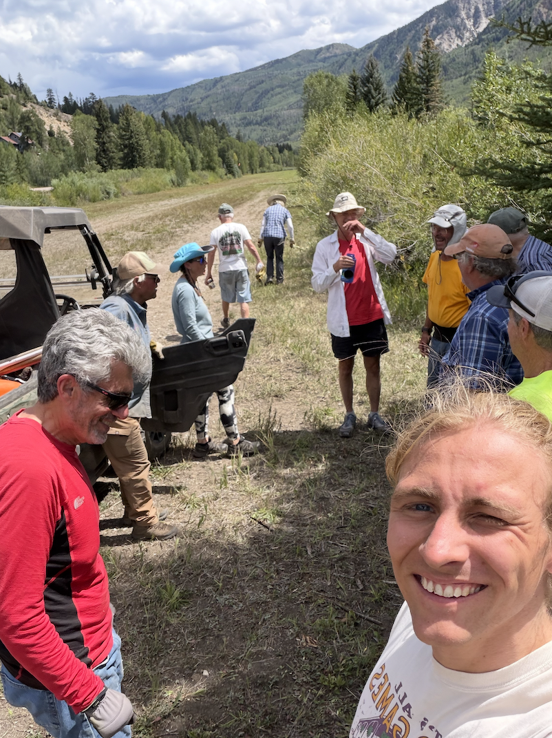A selfie on a grassy backcountry airstrip, with a group of volunteers and a utility vehicle behind.