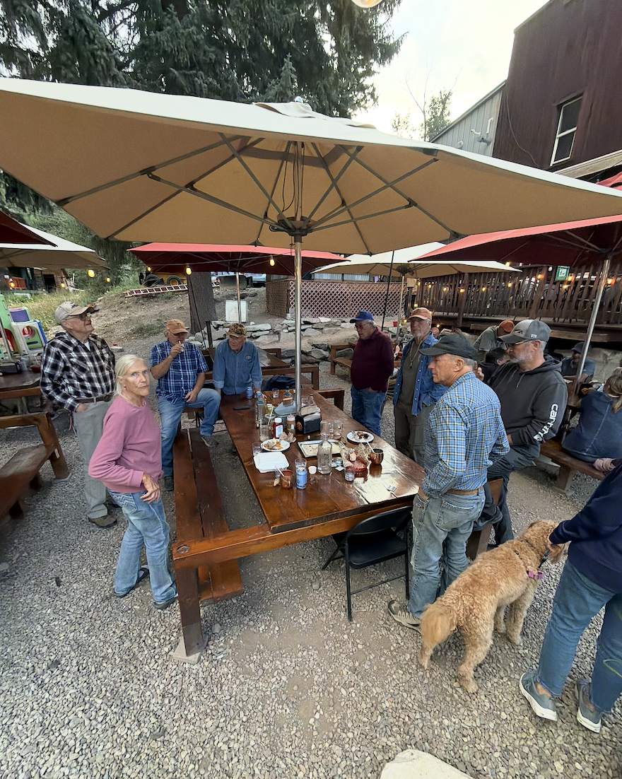 A group gathered at a long wooden table under a patio umbrella outside a restaurant, with a dog nearby.