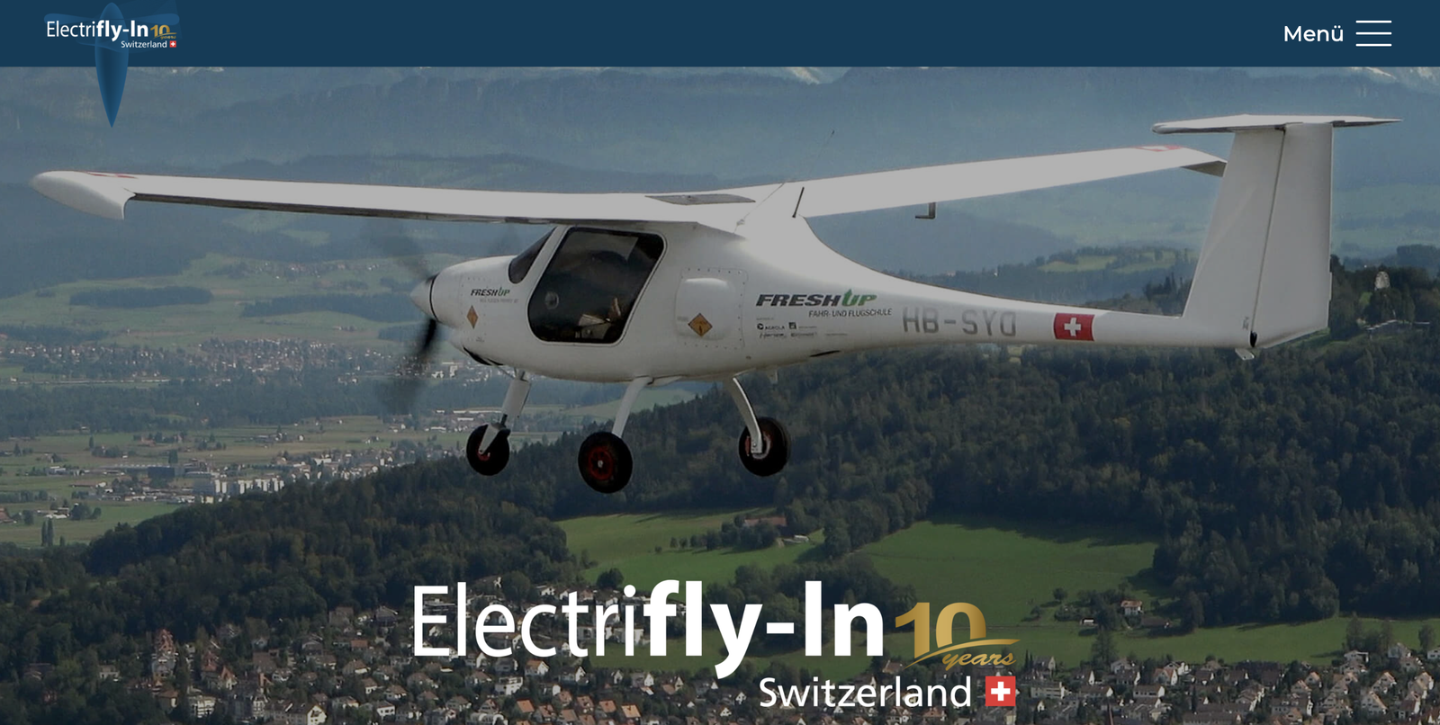A white electric light aircraft, registration HB-SYD, flying over green Swiss countryside, with the Electrifly-In 10 years Switzerland logo.