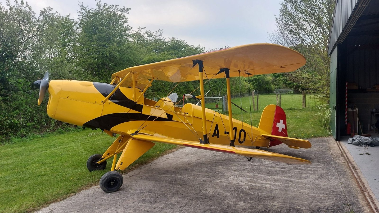 A yellow Bücker Jungmann biplane marked A-10, with a Swiss cross on the tail, parked outside a hangar.