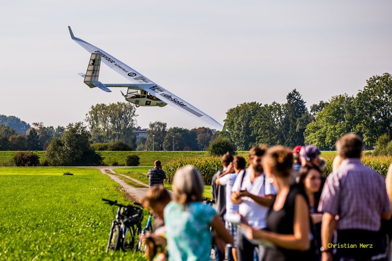 A long-winged solar-powered aircraft flying low over a grass field as spectators watch.