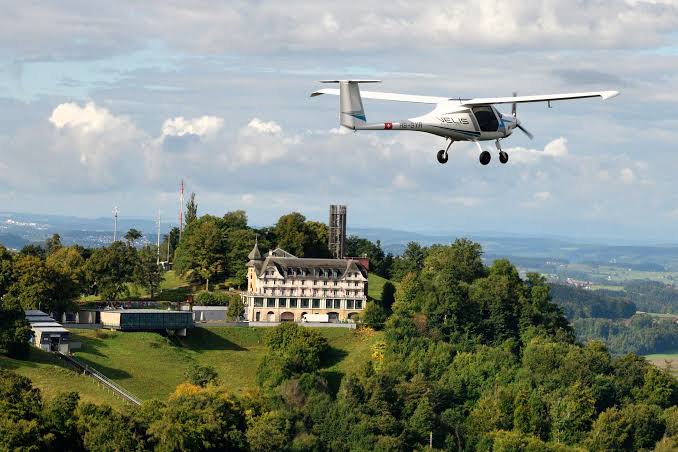 A white electric two-seat aircraft flying over a green hillside, with a large historic building below.