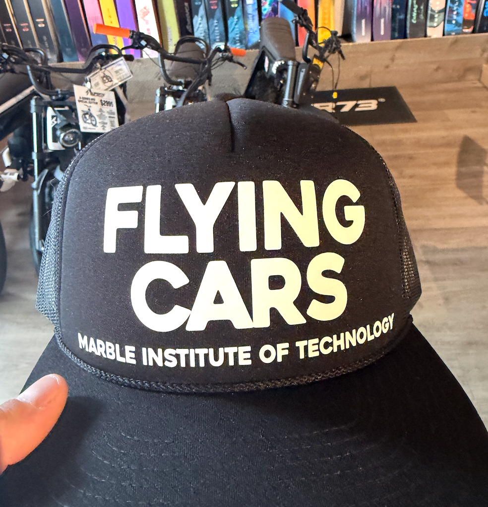 A black trucker hat that reads Flying Cars, Marble Institute of Technology.