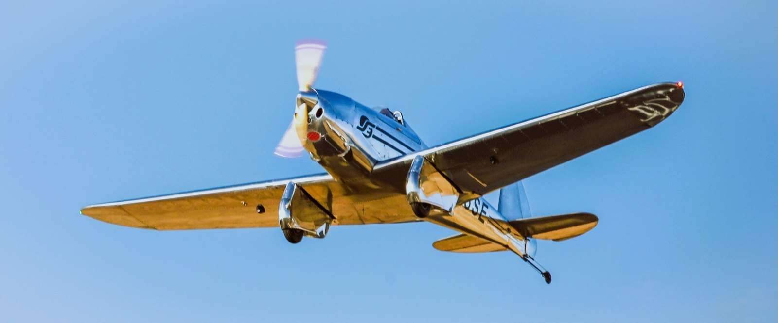 A polished silver single-engine airplane flying overhead against a blue sky.