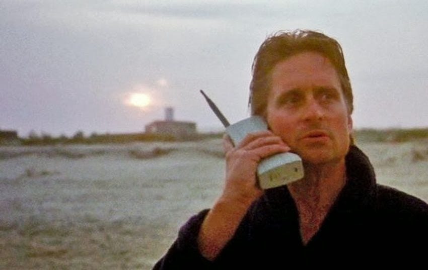 A film still of a man in a dark robe on a beach at dawn, talking on a large 1980s brick mobile phone.
