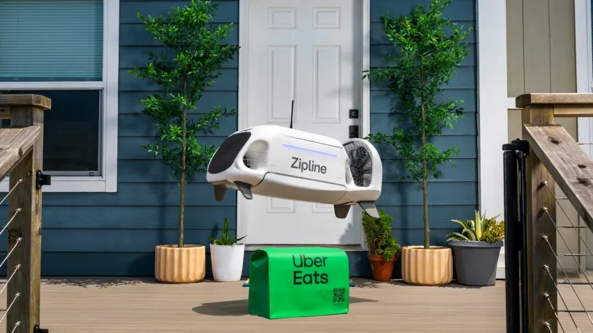A white Zipline delivery drone hovering over a green Uber Eats bag on the porch of a house.