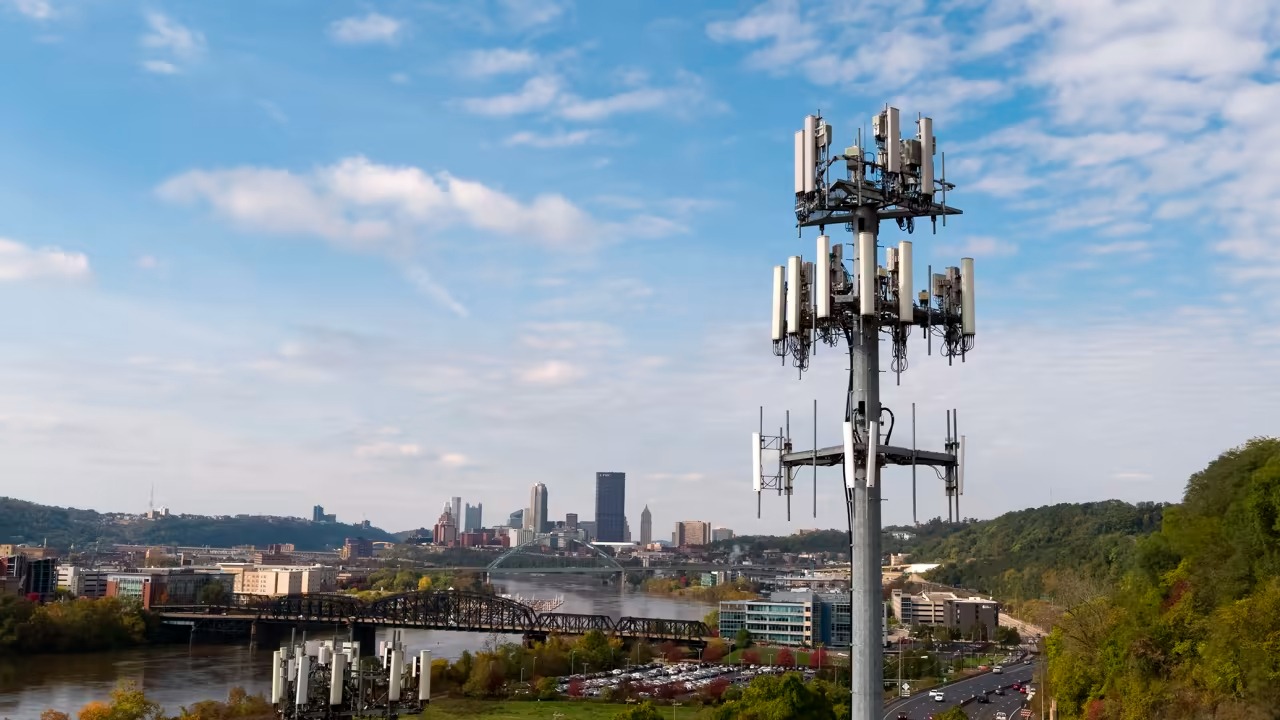 A cell tower covered in antennas, with the Pittsburgh skyline and a river behind it.
