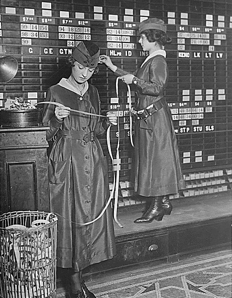 A black and white photo of two women in uniform posting stock prices by hand on a large quotation board, one reading ticker tape.