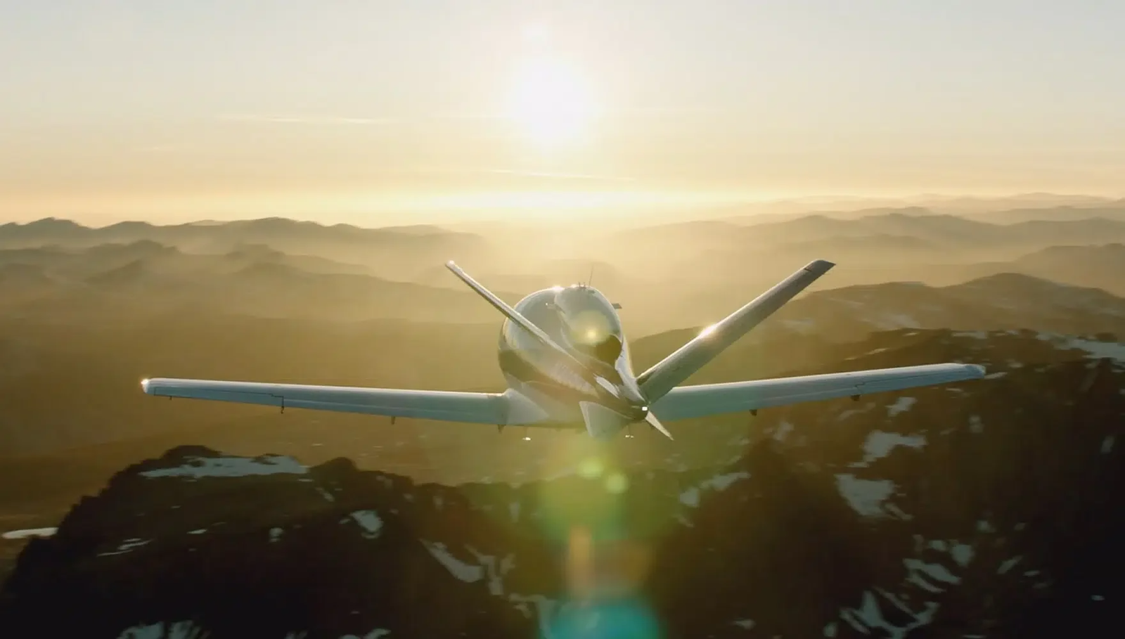 A single-engine airplane seen from behind, flying toward the setting sun over snowy mountain ridges.